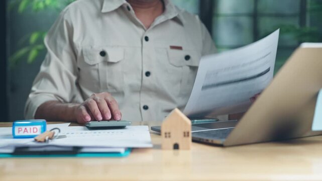 Senior man reviews utility bills calculates home mortgage expenses at wooden desk beside laptop, keys, rubber stamp, miniature wooden house model.