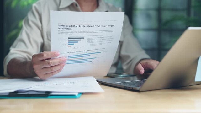Financial analyst reviewing printed investment report sheets beside laptop computer on office desk, analyzing shareholder float data corporate revenue metrics carefully indoors.