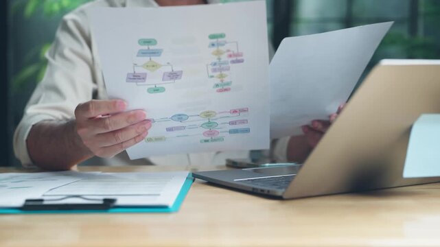Focused businessman reviewing printed flowchart documents beside open laptop on wooden table in office setting, analyzing corporate project management diagrams, workflow procedures, planning.