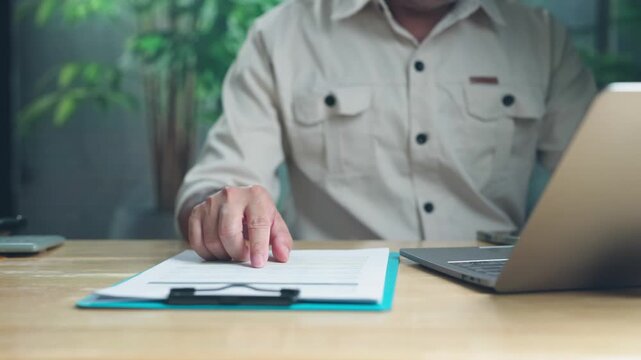 Focused businessman pointing index finger at financial document on wooden desk, reviewing accounting report data beside laptop computer in contemporary workspace office.