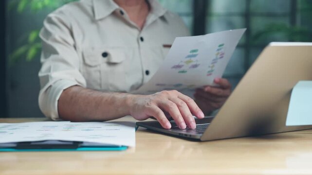 Focused engineer reviews printed process flowcharts holding documentation before typing on laptop keyboard at wooden desk inside contemporary office workspace.