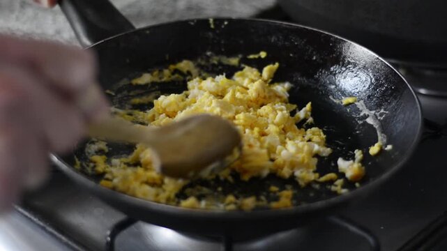 Close up of hand stirring creamy scrambled eggs in a frying pan with a wooden spoon