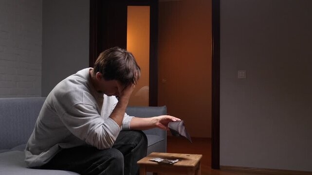 Young man sitting on a sofa at home, holding an empty wallet and looking upset, depicting concepts of poverty, debt, and unemployment in a gloomy interior