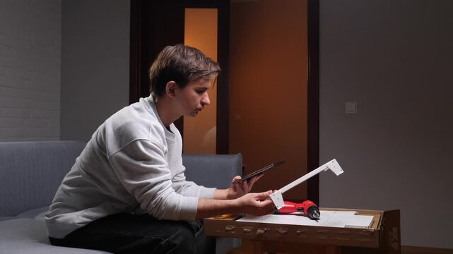 Young man assembling new flat pack furniture at home, focusing on digital instructions displayed on a tablet, engaging in a diy project for home improvement