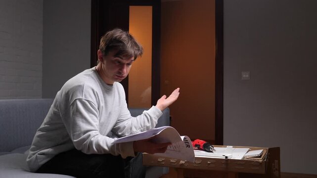 Man feeling confused and frustrated while reading complex instruction manual, attempting to assemble new flat pack furniture at home in his living room