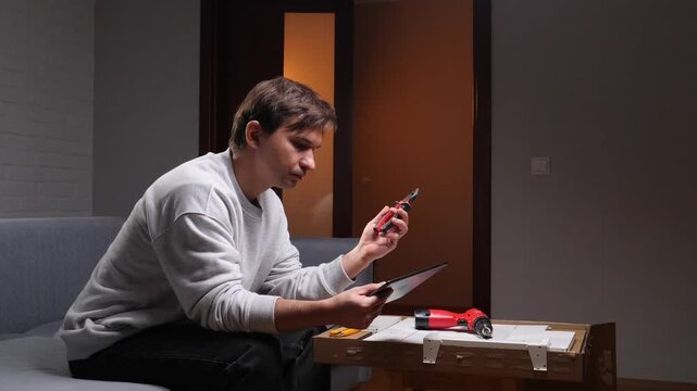 Man on sofa intently following tablet tutorial while holding pliers, assembling furniture at homediy project, focused, learning new skills with online instructions
