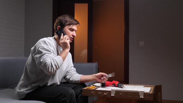 Man at home talking on a mobile phone for assistance while attempting to assemble flat pack furniture, holding a piece and looking at an instruction manual