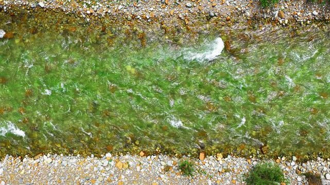Top down aerial view captures clear emerald water flowing rapidly over smooth pebbles along a scenic mountain stream.