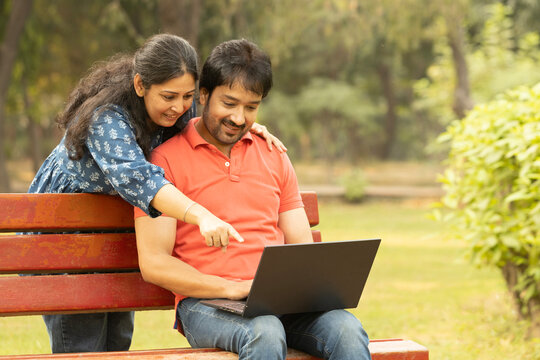 Indian happy couple working on laptop and doing online payment in park. Concept internet banking