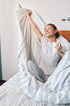 Happy young woman stretching in bed under blanket holding white mug. Morning waking up concept. Cozy home lifestyle, good mood and restful sleep.