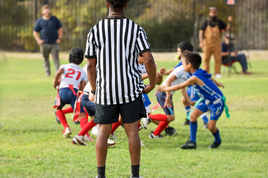 A football referee in a black and white striped uniform stands on a grass field, watching young children play an active game of youth flag football.