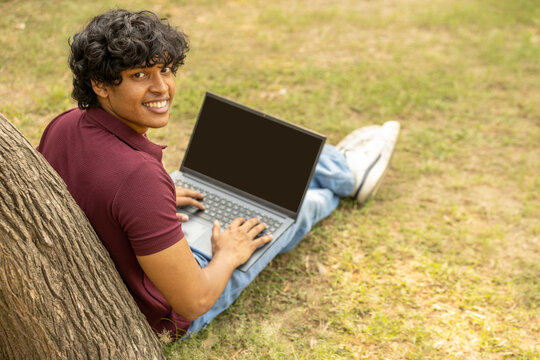 Indian happy college student boy showing laptop screen at park for social media online marketing. Black screen, digital technology and social network advertising for brand promotions