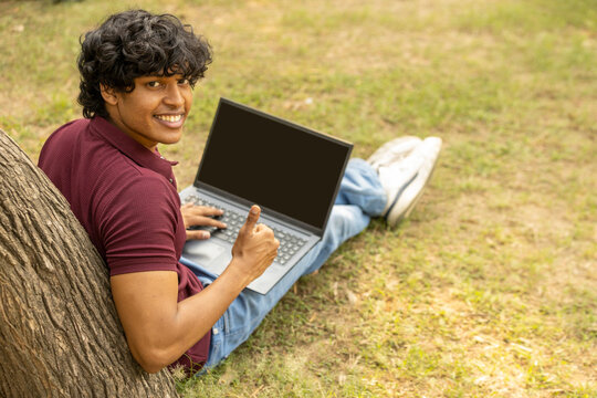 Indian happy college student boy showing thumbs up with laptop screen at park for social media online marketing. Black screen, digital technology and social network advertising for brand promotions