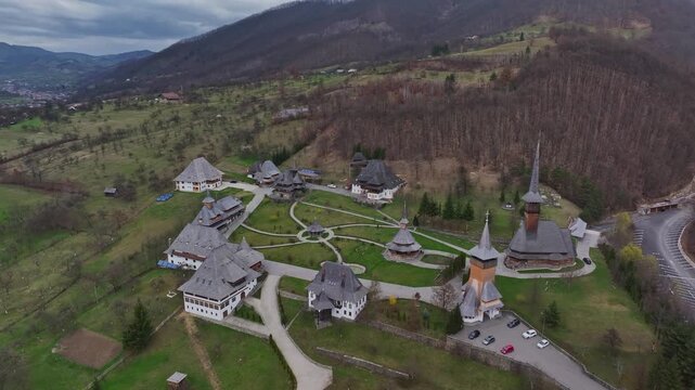 Aerial drone footage revealing the B&acirc;rsana Monastic Complex with its iconic wooden Orthodox church, towering wooden spire, traditional roofs, and surrounding green hills. Located in Maramureș County