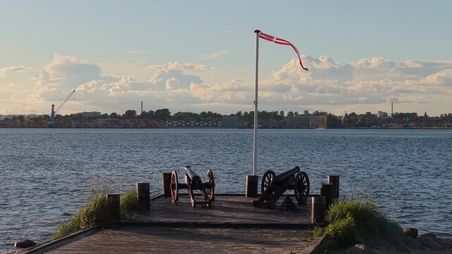 Footage of two historic black cast-iron cannons on wooden carriages facing Fredericia across the Little Belt in Middelfart, Denmark, at the Tre&aring;rskrigen memorial waterfront.