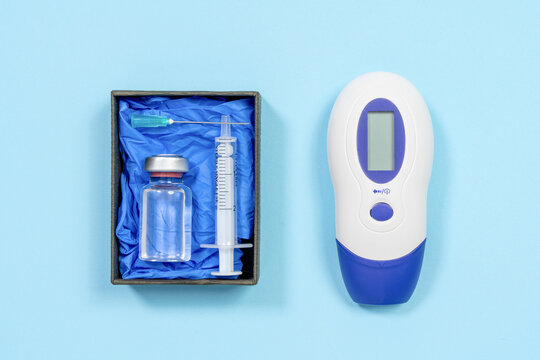 A boxed glass ampoule with syringe and needle rests beside a handheld infrared device with blank digital display on a blue surface, a concept of seasonal illness monitoring at home.