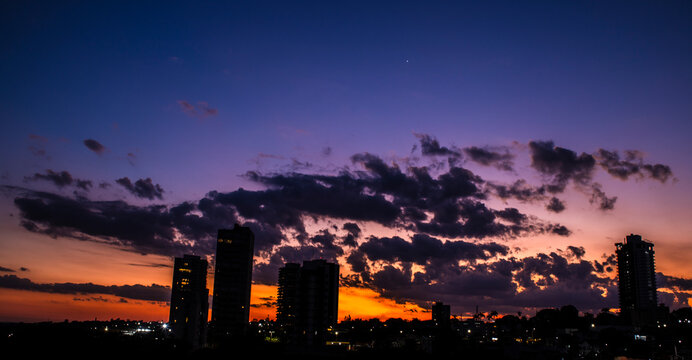 A breathtaking city skyline silhouette under a dramatic twilight sky filled with deep purple clouds and a vibrant orange sunset glow, featuring a single bright star or planet visible