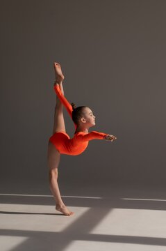 A little Caucasian girl gymnast in an orange leotard demonstrates her skills on a white background. 