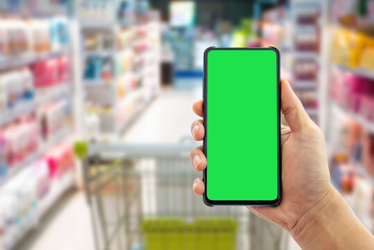 Person holding smartphone with chroma key green screen for template mockup in blurred grocery store. Retail and online shopping concept. Human-created, not AI.