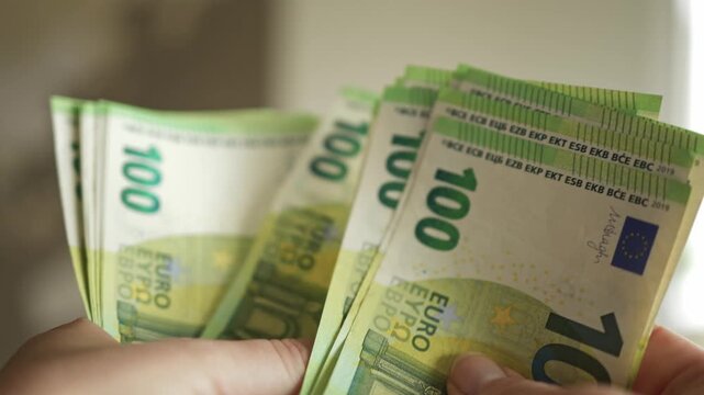 Close-up of hands displaying several 100 euro banknotes, highlighting the intricate details of the currency with a softly blurred background