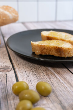 Golden-brown bread slices rest on a black plate. Green olives surround the plate on rustic wood. Fresh, appetizing, and simple. Perfect for food blogs or restaurant menus.