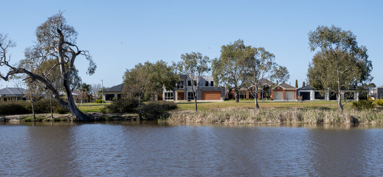 A calm view across the Wimmera River with contemporary homes or residential houses set back from the riverbank, framed by leaning native eucalyptus trees and reeds. Waterfront housing in regional town