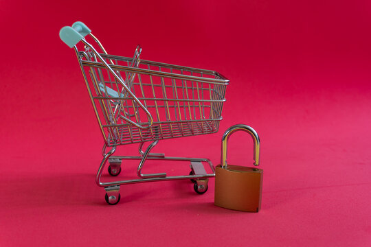 Tiny shopping cart stands beside a padlock on red background. Symbolizes blocked access or online shopping restrictions. Suggests security, limited availability, or payment issues