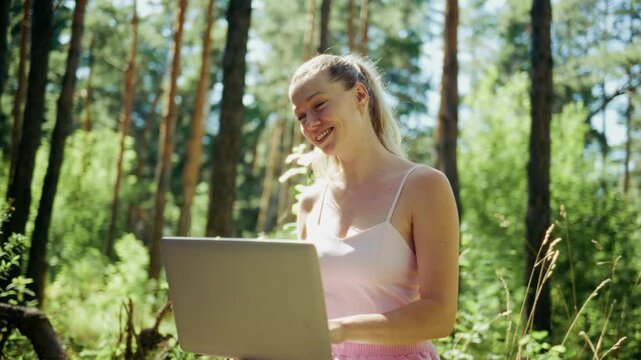 Young woman smiles in sunlit forest, laptop open. She breathes deeply, eyes closed, enjoying nature. Peaceful moment of work or relaxation outdoors. Ideal for wellness, remote work