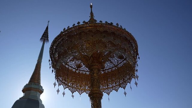 An intricate golden metal umbrella stands against a clear blue sky at a northern Thai temple. The sacred pagoda shines brightly in the sunny architectural background.