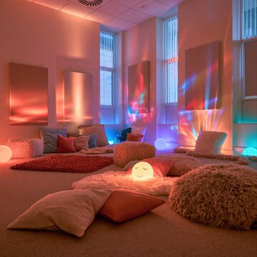 Color-changing light installation in meditative space with stacked round cushions, abstract shapes, and pink-blue neon gradients throughout.