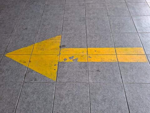 A large weathered yellow directional arrow painted on a gray tiled pavement, pointing to the left and symbolizing guidance, direction, navigation, or movement.