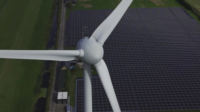 Aerial view of a large wind turbine standing next to an extensive solar farm with rows of panels in the rural landscape of 't Zand, North Holland, Netherlands.
