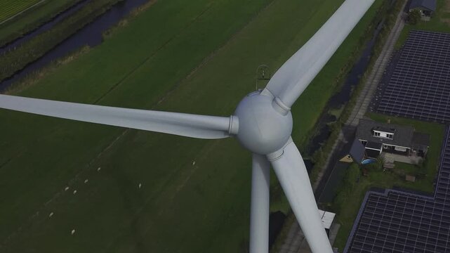 Aerial view of a wind turbine blade rotating near a field of solar panels and a residential house in the countryside of 't Zand, North Holland, Netherlands.