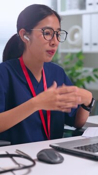 Asian woman healthcare worker nurse medical professional wearing eyeglass earphone headset using computer laptop at office desk, medical worker making gesture in navy uniform Vertical
