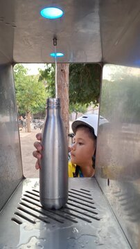 A boy wearing a bicycle helmet fills a metal water bottle at an outdoor drinking fountain in a tree-lined park, Vertical