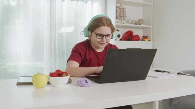 Child doing homework with a laptop. Child studying beside a notebook and pencil. Studious child using technology for homework. Focused child working at a home desk.