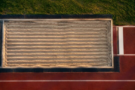 Raked runway of a long jump pit patterned with clean parallel furrows in pale dry sand and damp grains along the grooves