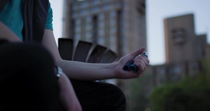 Man holding a pen in a city park during sunset while sitting on a bench with tall buildings in the background
