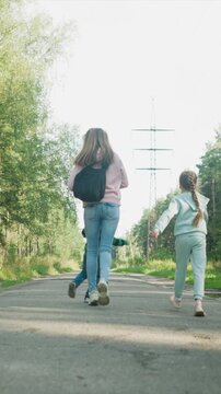 Family walks together down tree lined road. young mother with long hair wearing pink hoodie and carrying black backpack is followed by her son in green striped shirt and daughter dressed all