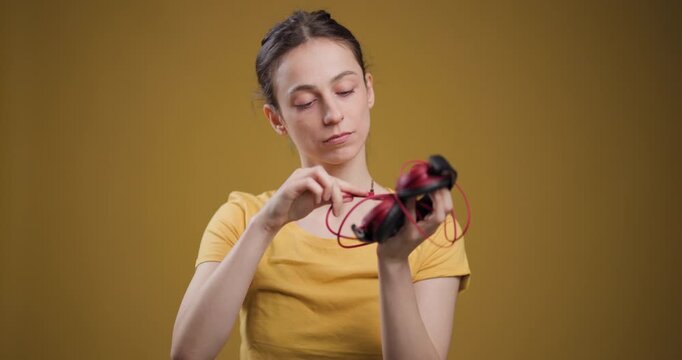 Young woman in yellow t-shirt untangling headphones against a mustard background