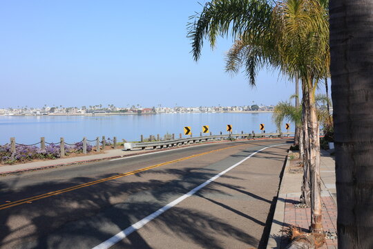 Yellow chevron signs and a metal guardrail guide traffic along the asphalt street next to the scenic Mission Bay and distant shoreline, San Diego, California