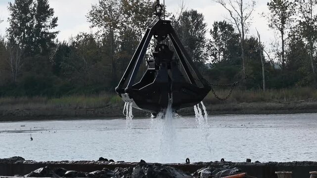 Heavy steel clamshell bucket hangs over a barge and releases wet dredged mud back onto the deck beside a quiet riverbank.