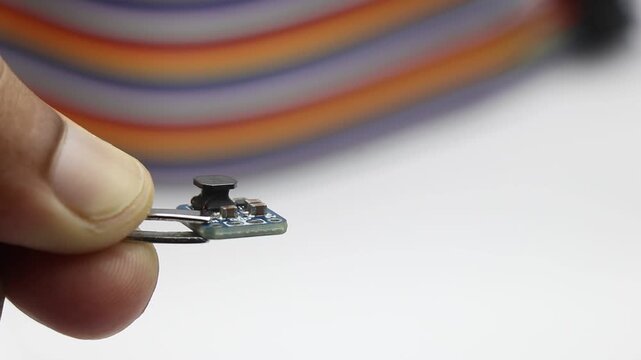 Tweezers holding small power regulating board with side view of transformer on ribbon cable background