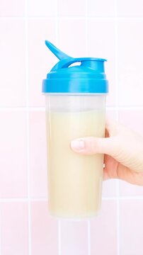 Female hand holds shaker with protein shake against background of pink tiles.