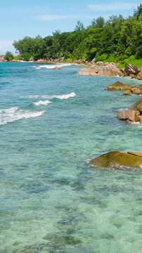 Waves break on rocky shores along a coastline covered in lush tropical greenery. La Digue, Seychelles.