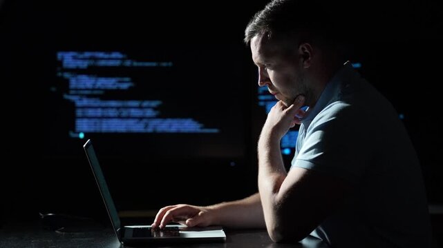 Programmer coding on a laptop computer. Programmer working with code on multiple screens. Male programmer typing at a dark workstation. Focused programmer developing software at a laptop.