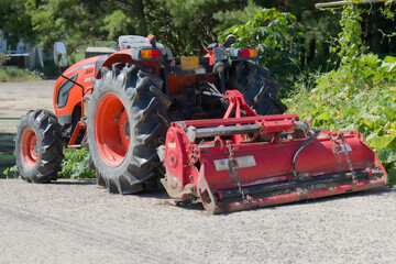 Orange Compact Tractor with Red Rotary Tiller Attachment Parked on a Dirt Road in a Rural Setting © L.Z.C Photographer