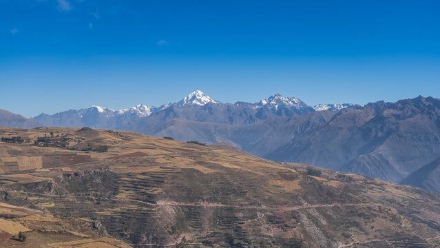 Beautiful high-altitude landscape. Andes mountain range with snow-capped peaks against a clear blue sky. There are rural houses, cultivated fields, and terraces in the valley. Peru. Copy space