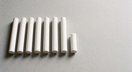 Fototapeta premium Row of blank white chalk sticks with one short broken piece on a light grey paper background with copy space