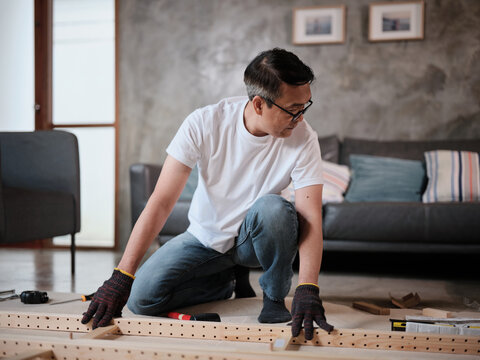 Asian mature man self-assembling wooden flat-pack furniture with DIY carpentry tools, following instruction manual, home improvement, craft installation, hobby workshop, handyman active lifestyle.